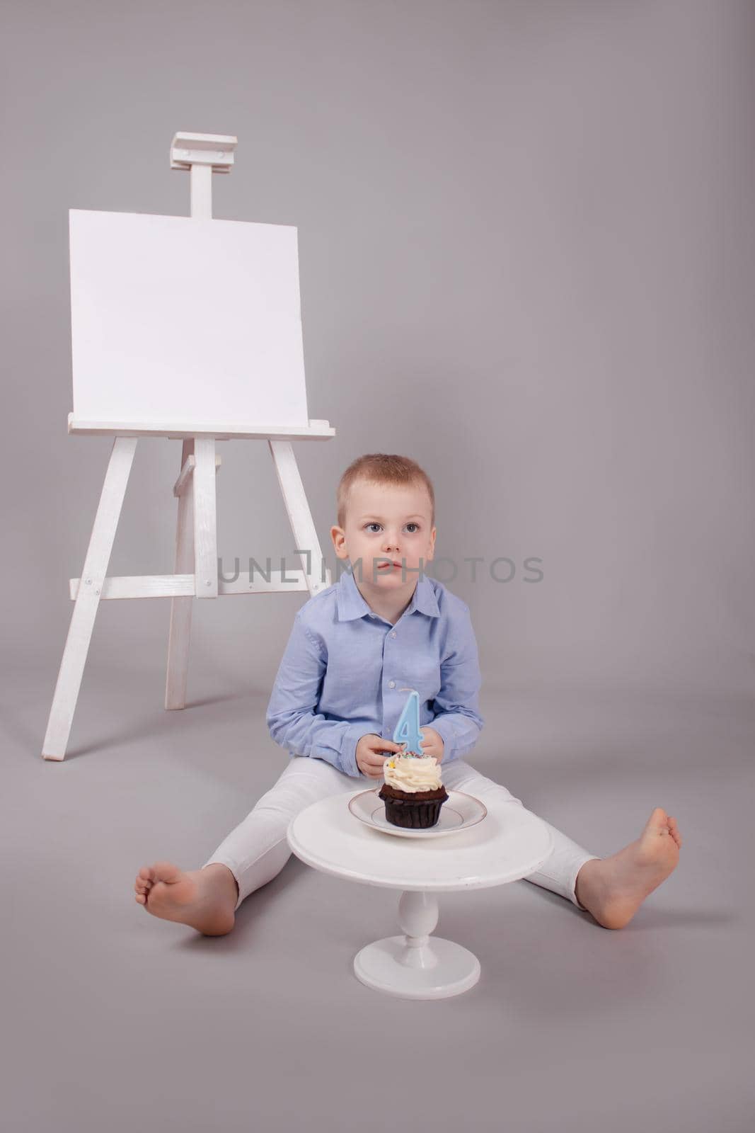 preschool boy in white pants and blue shirt on grey background near easel with mockup and cupcake with candle, shaped in number four 4. happy birthday. celebration. High quality photo