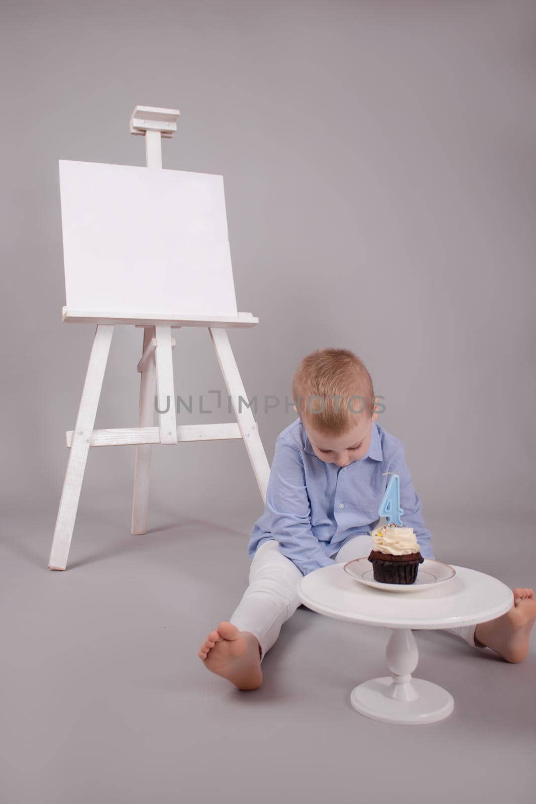 preschool boy in white pants and blue shirt on grey background near easel with mockup and cupcake with candle, shaped in number four 4. happy birthday. celebration by oliavesna