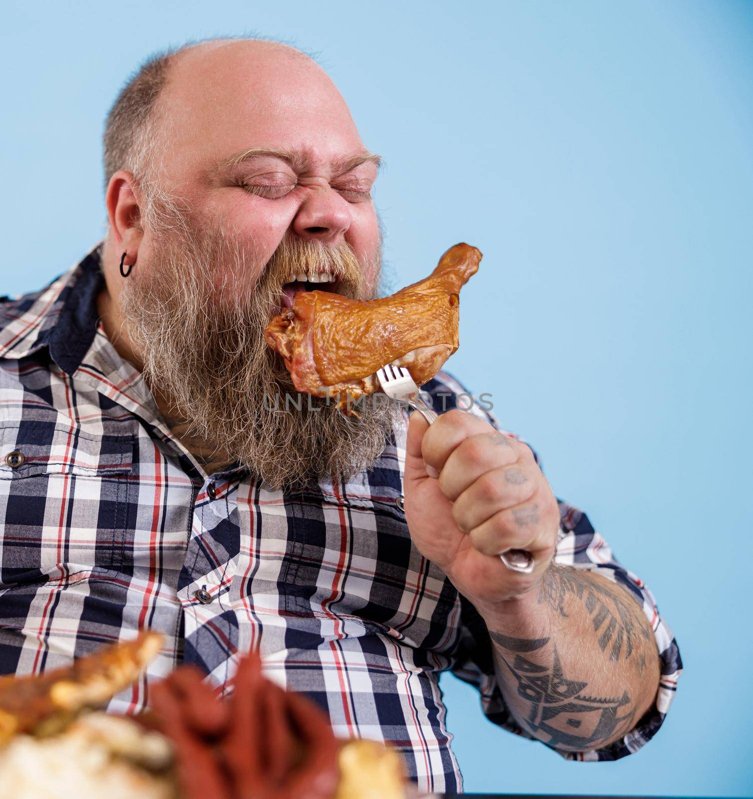Funny middle aged bearded man with overweight bites piece of smoked chicken leg sitting on light blue background in studio