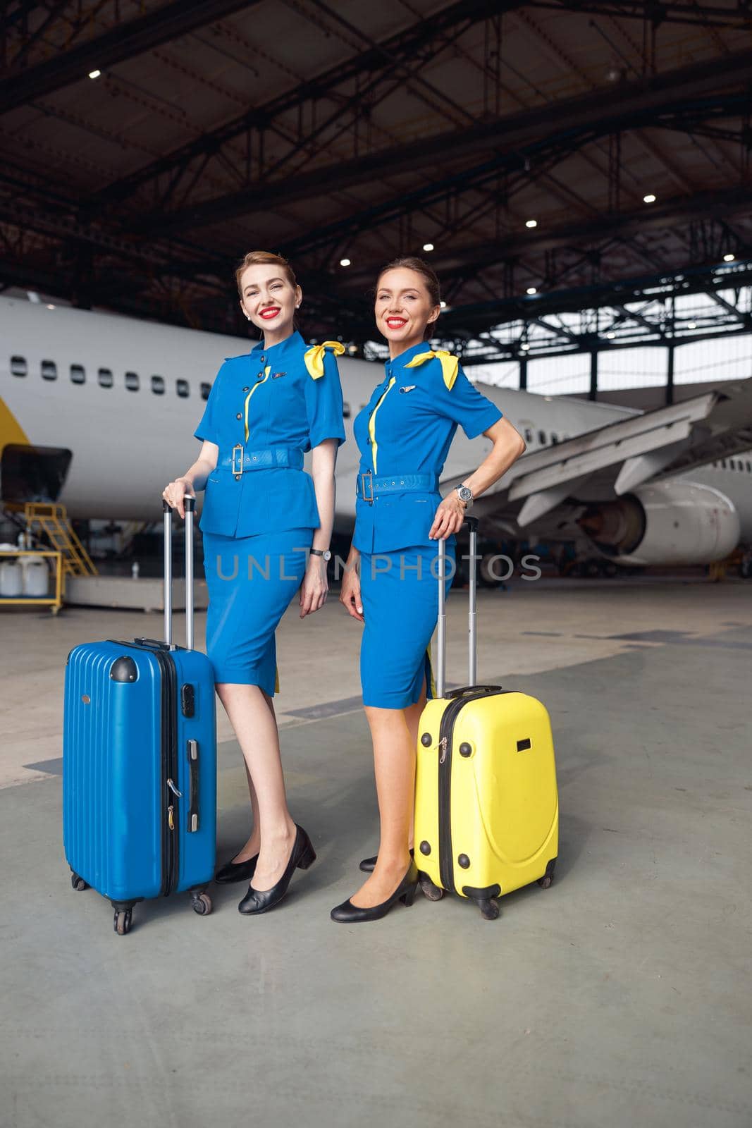 Full length shot of two smiling air stewardesses in bright blue uniform ready for flight, standing with their luggage in front of passenger aircraft in hangar at the airport. Occupation concept