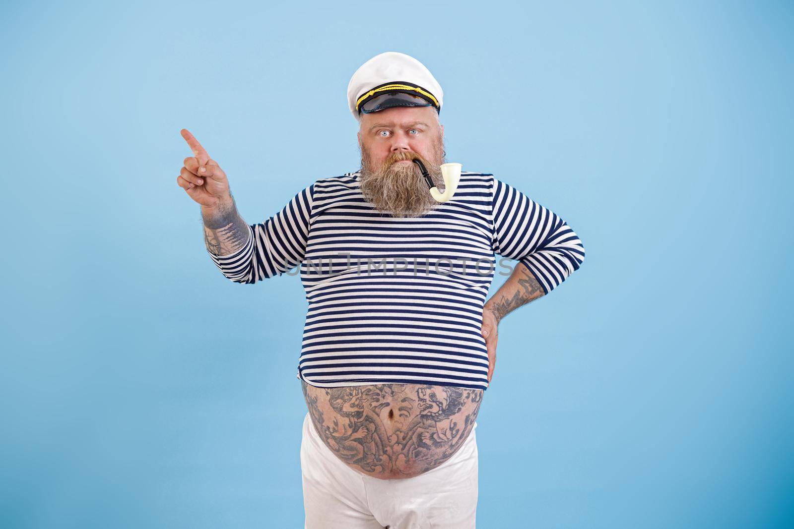 Plus size man with tattoos on large tummy in sailor suit with ancient smoking pipe points aside by finger on light blue background in studio