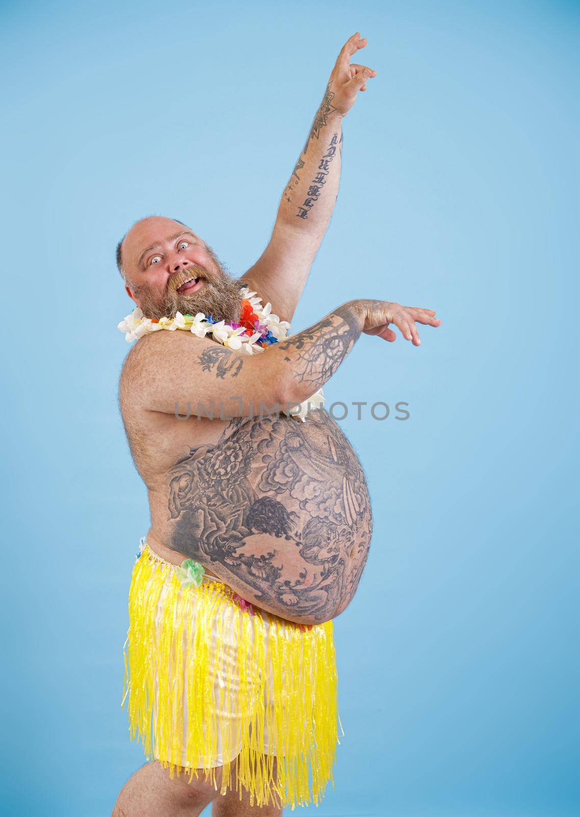Funny expressive bearded obese man wearing decorative yellow grass skirt and flowers garland dances on light blue background in studio