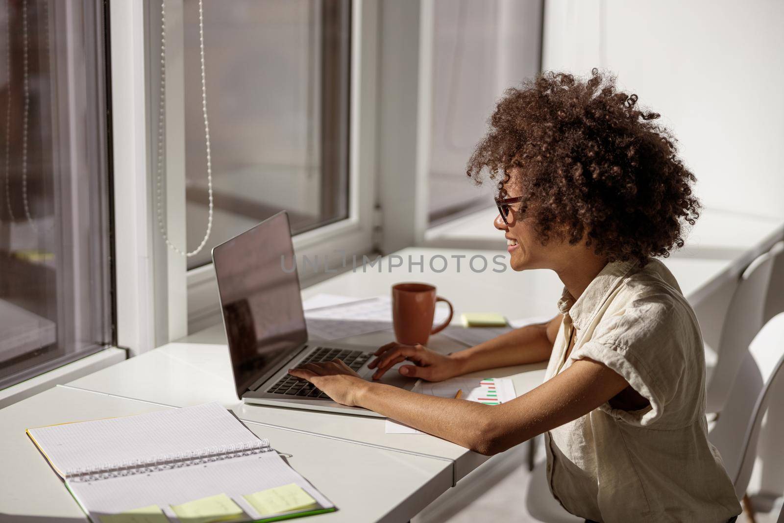 Smiling concentrated female administrative typing messages on laptop while sitting near the window in the office