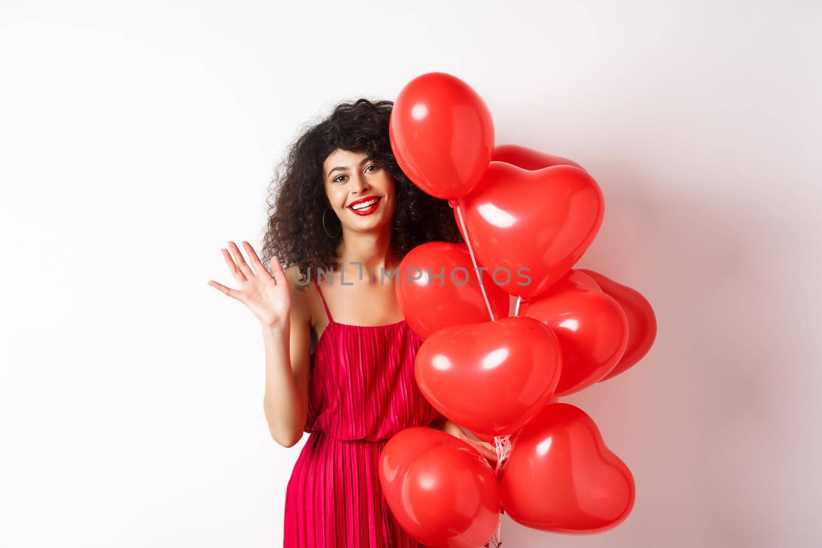 Beautiful lady with curly hair, standing near valentines holiday balloons and waving hand, saying hello, standing against white background by Benzoix