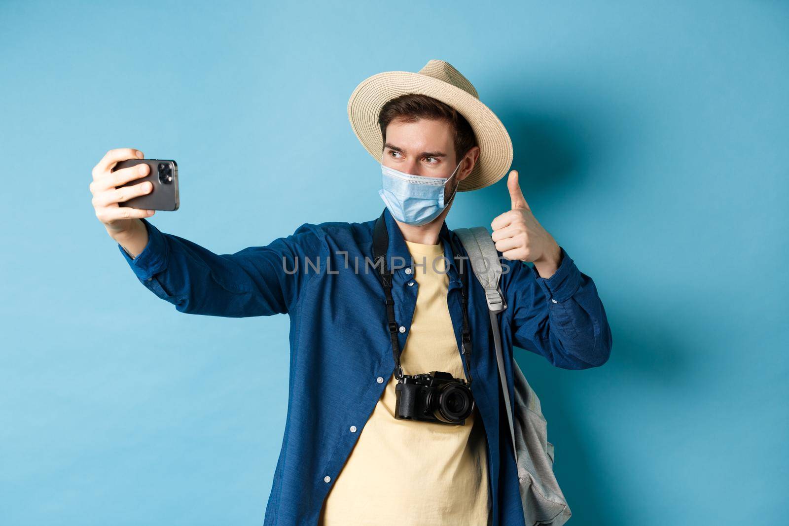 Covid-19, pandemic and travel concept. Happy male tourist taking pictures on vacation, make selfie with thumb up, recommending hotel, standing on blue background.