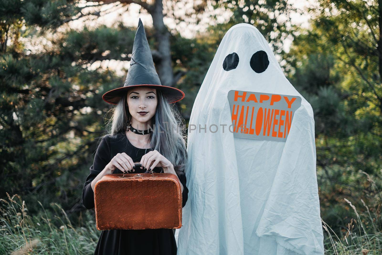 White ghost with empty poster and witch with suitcase walking in autumn forest, free space. Halloween theme