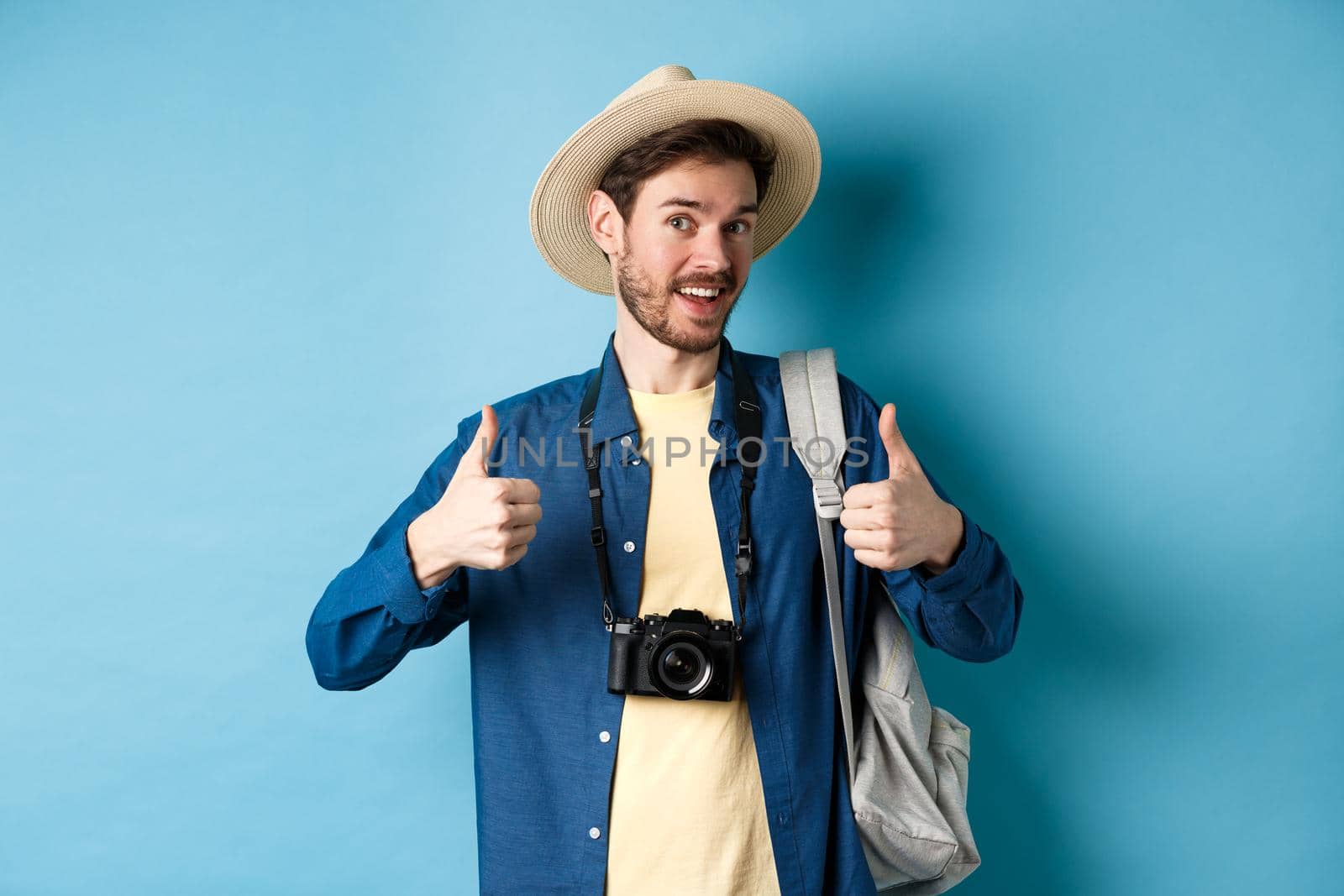 Happy smiling tourist say yes and show thumbs up, going on summer vacation, holding backpack and photo camera, blue background.