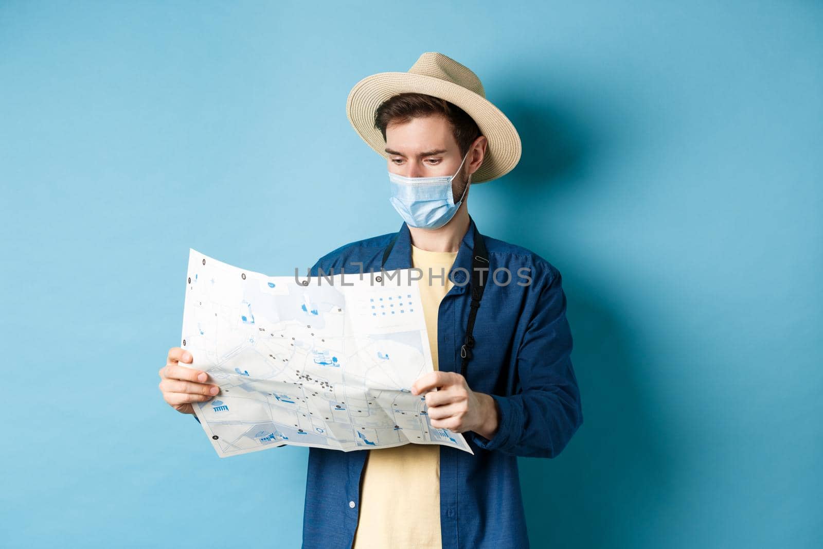 Covid-19, pandemic and travel concept. Tourist looking at map with sightseeing on vacation, wearing summer hat and medical mask from coronavirus, blue background.