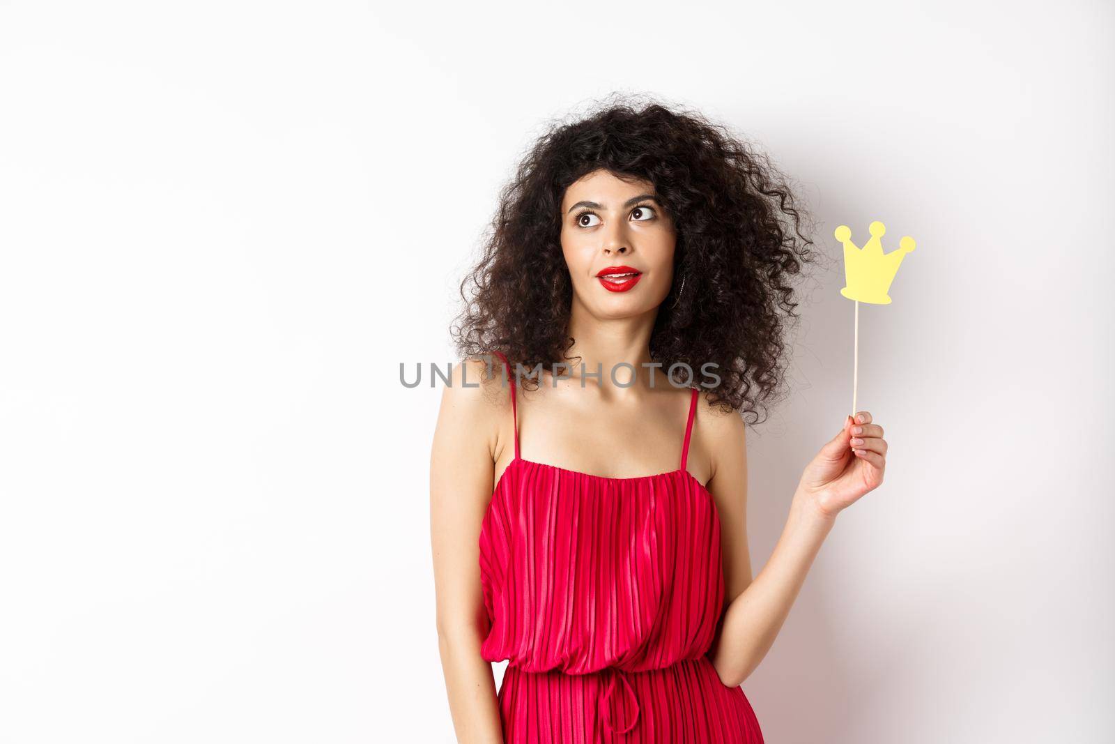 Stylish woman with curly hair in red dress, holding queen crown on stick and looking aside, standing on white background.