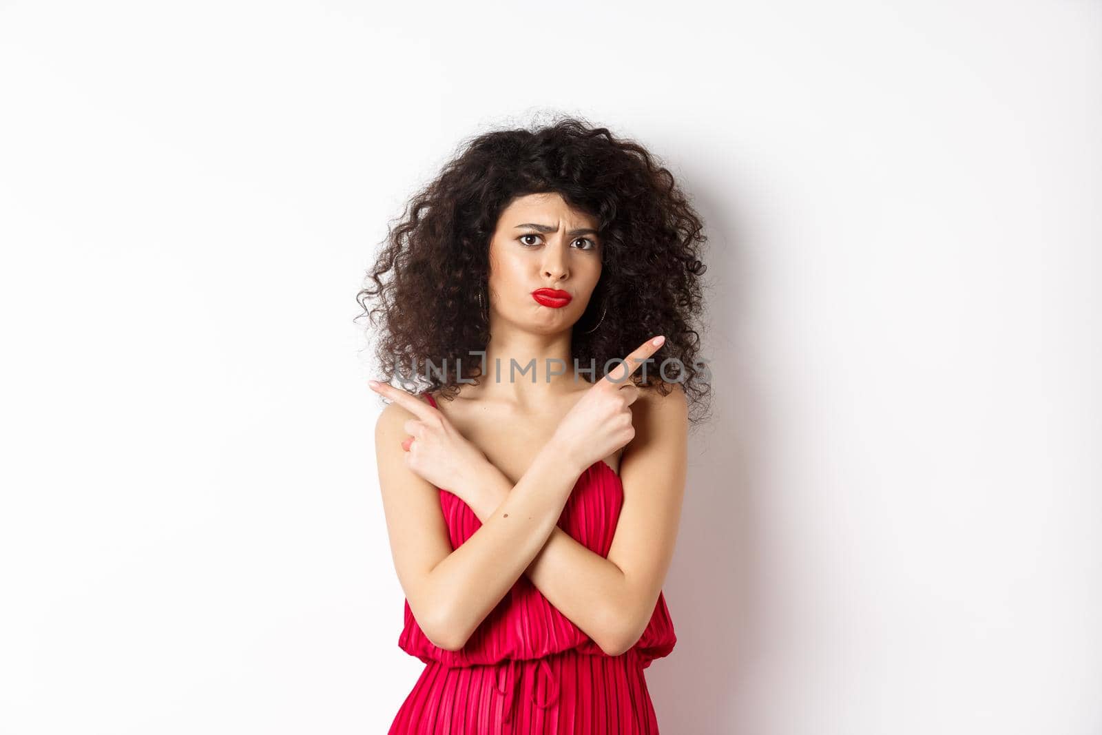 Doubtful and sad young woman in red dress, pointing fingers sideways and sulking, cant decide between products, need help with choice, standing over white background.