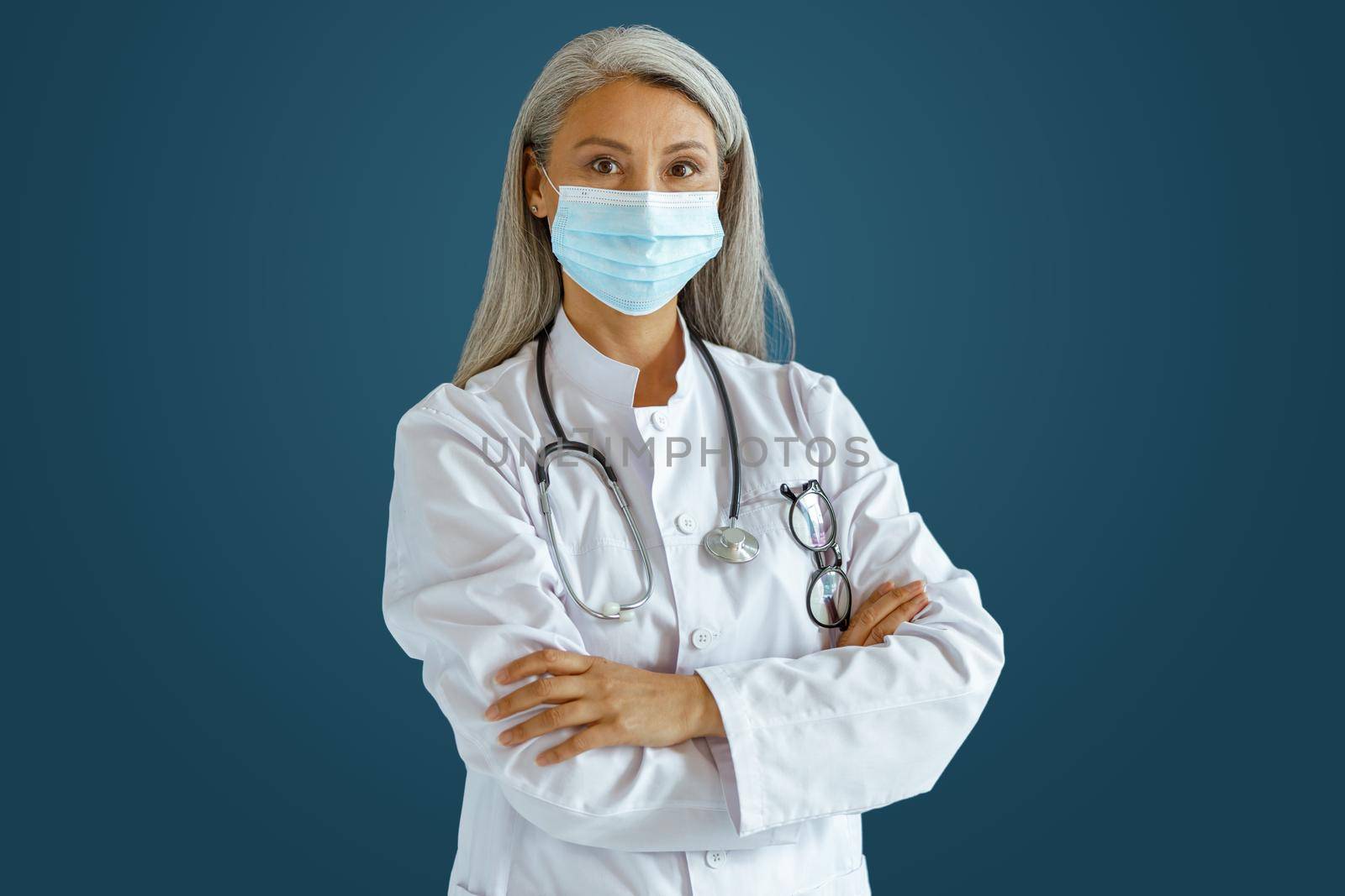 Grey haired Asian female doctor in white robe with medical mask and stethoscope stands on blue background in studio. Professional medical staff