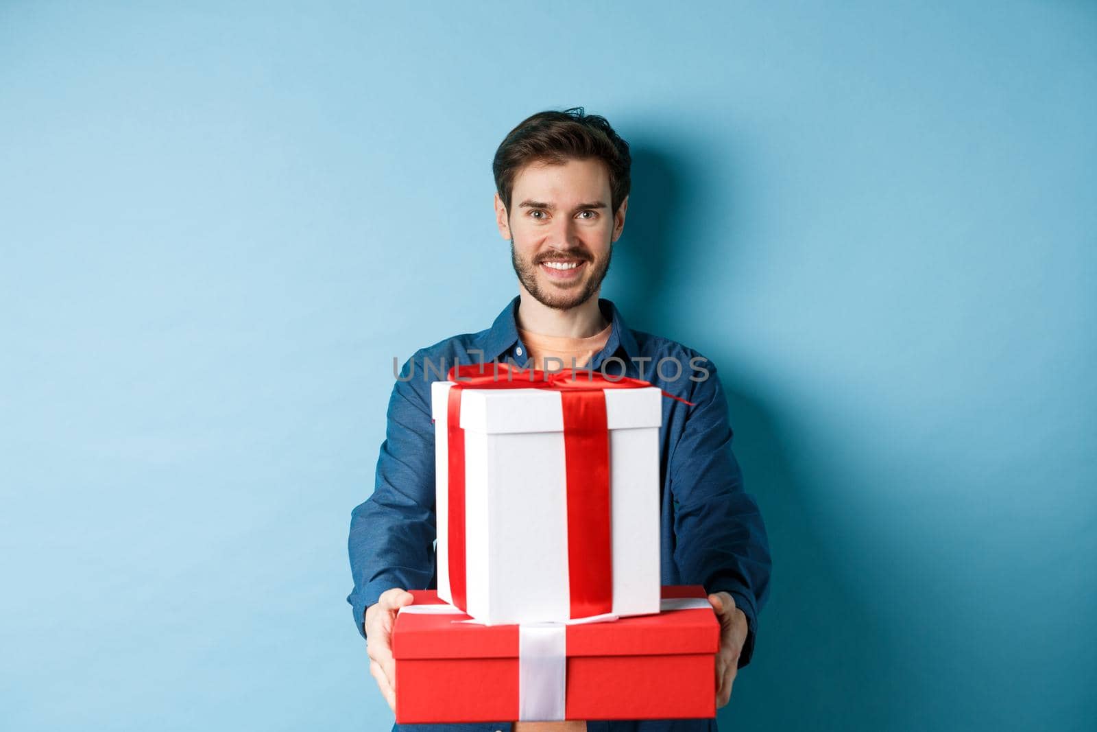 Happy valentines day. Handsome man giving girlfriend presents, holding gift boxes and smiling, standing over blue background.