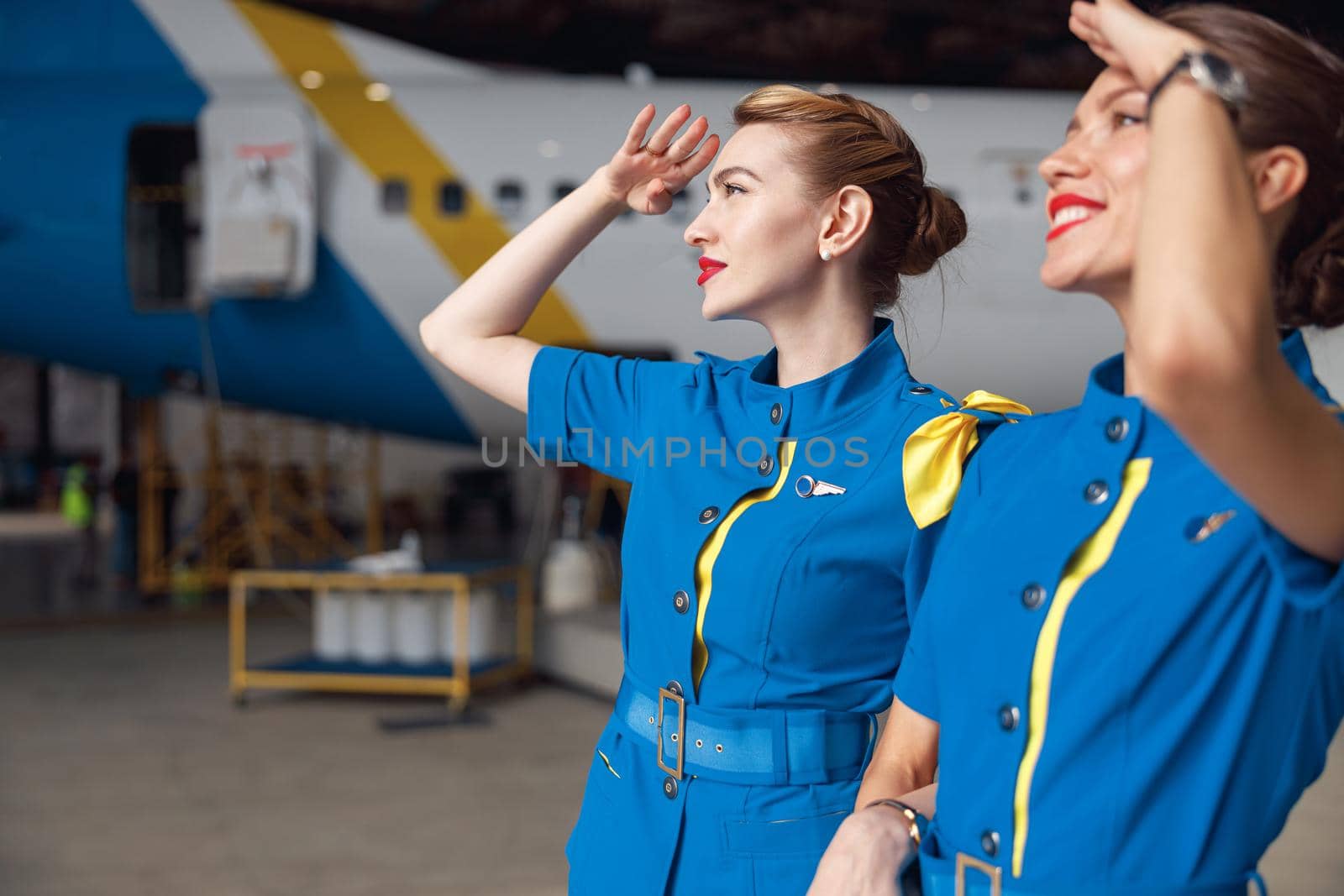 Portrait of two beautiful air stewardesses in stylish blue uniform smiling while looking up in the sky, standing together in front of passenger aircraft in hangar at the airport by Yaroslav_astakhov