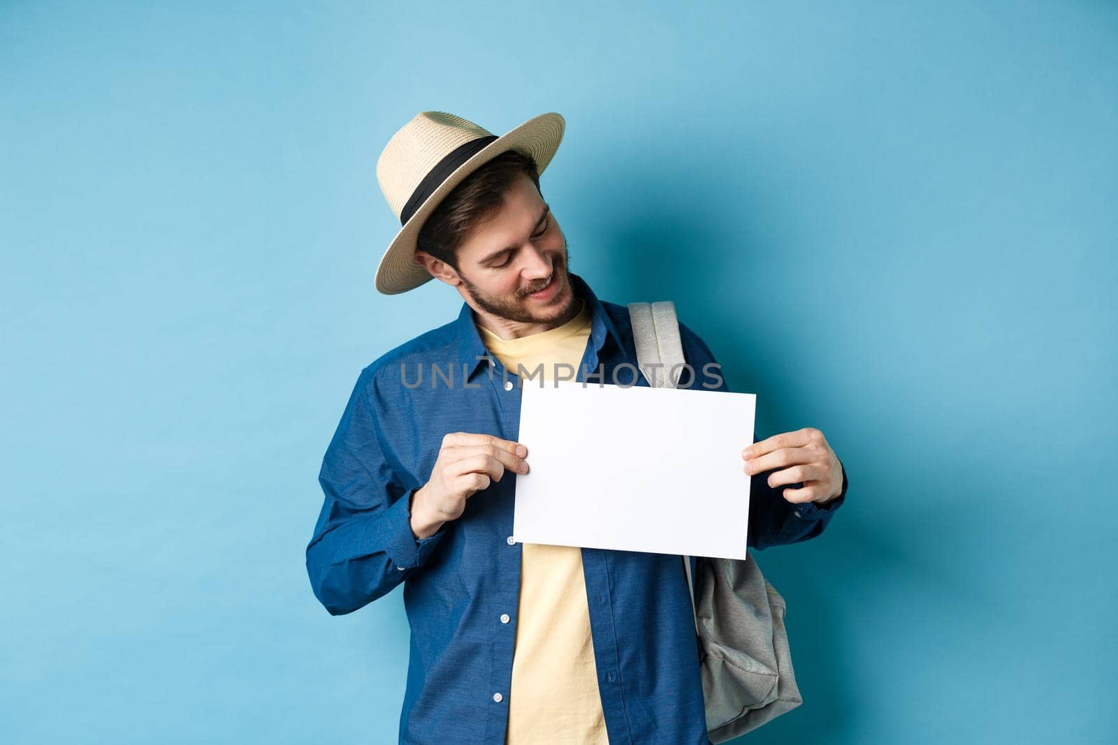 Happy tourist in straw hat smiling and looking at piece of paper for your logo, standing on blue background by Benzoix