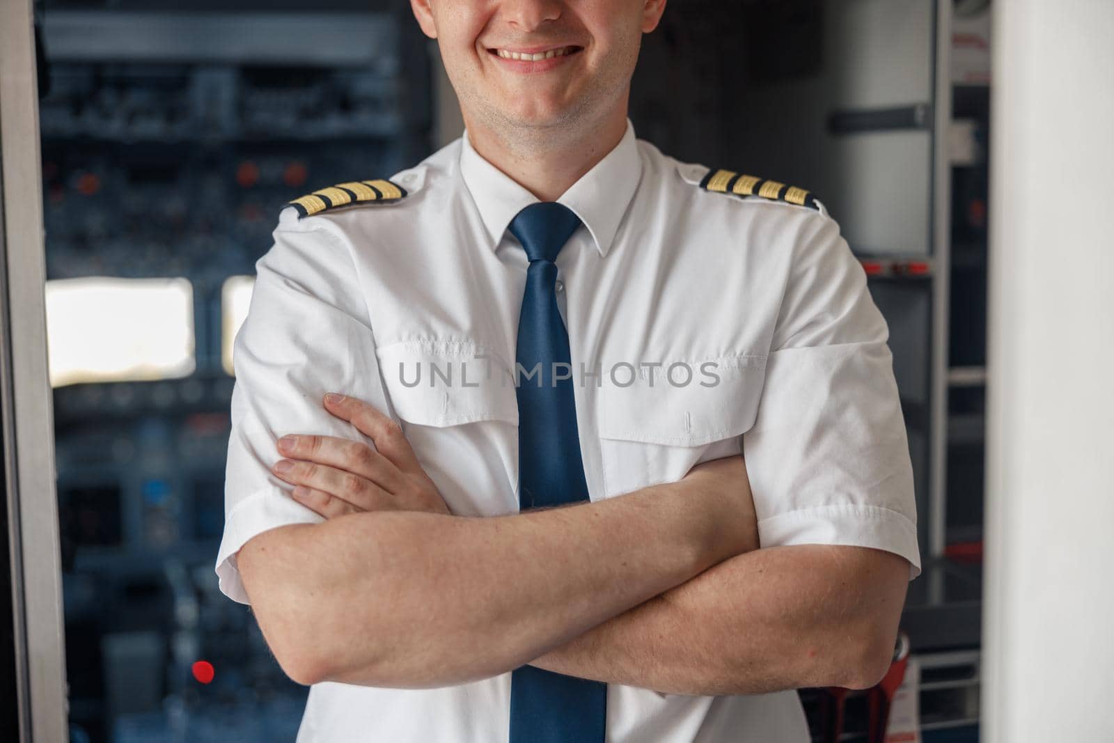Cropped portrait of male pilot in uniform keeping arms crossed and smiling while posing, standing inside of the airplane. Transportation, aircrew concept