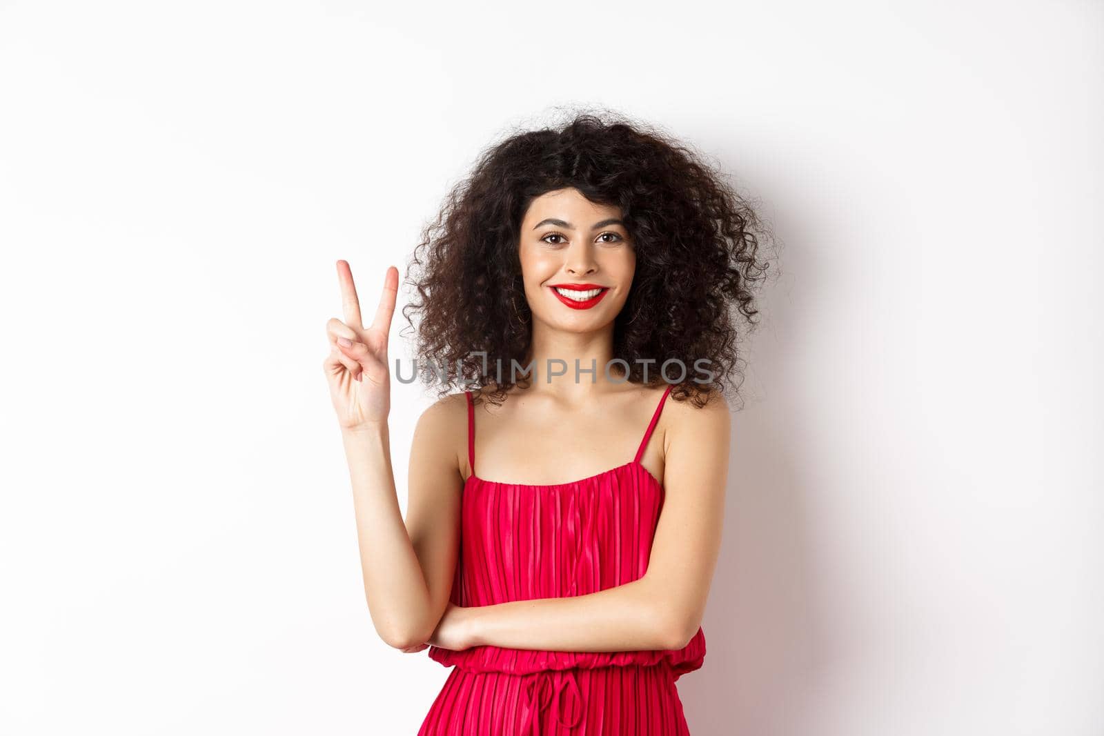 Attractive female model in red dress and makeup, showing number two and smiling, standing over white background.
