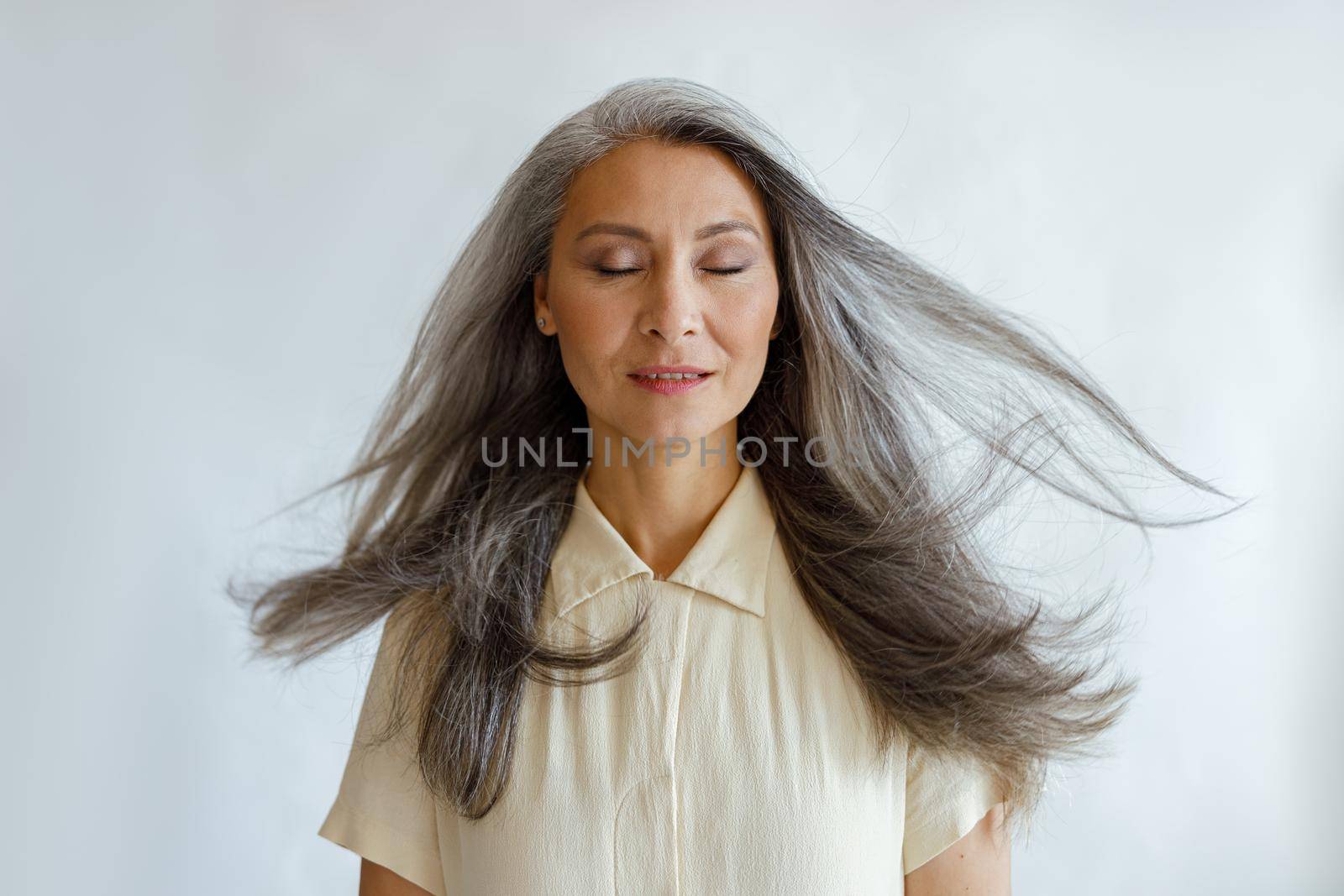 Pretty tranquil Asian woman with flying silver hair stands on light grey background in studio. Mature beauty lifestyle