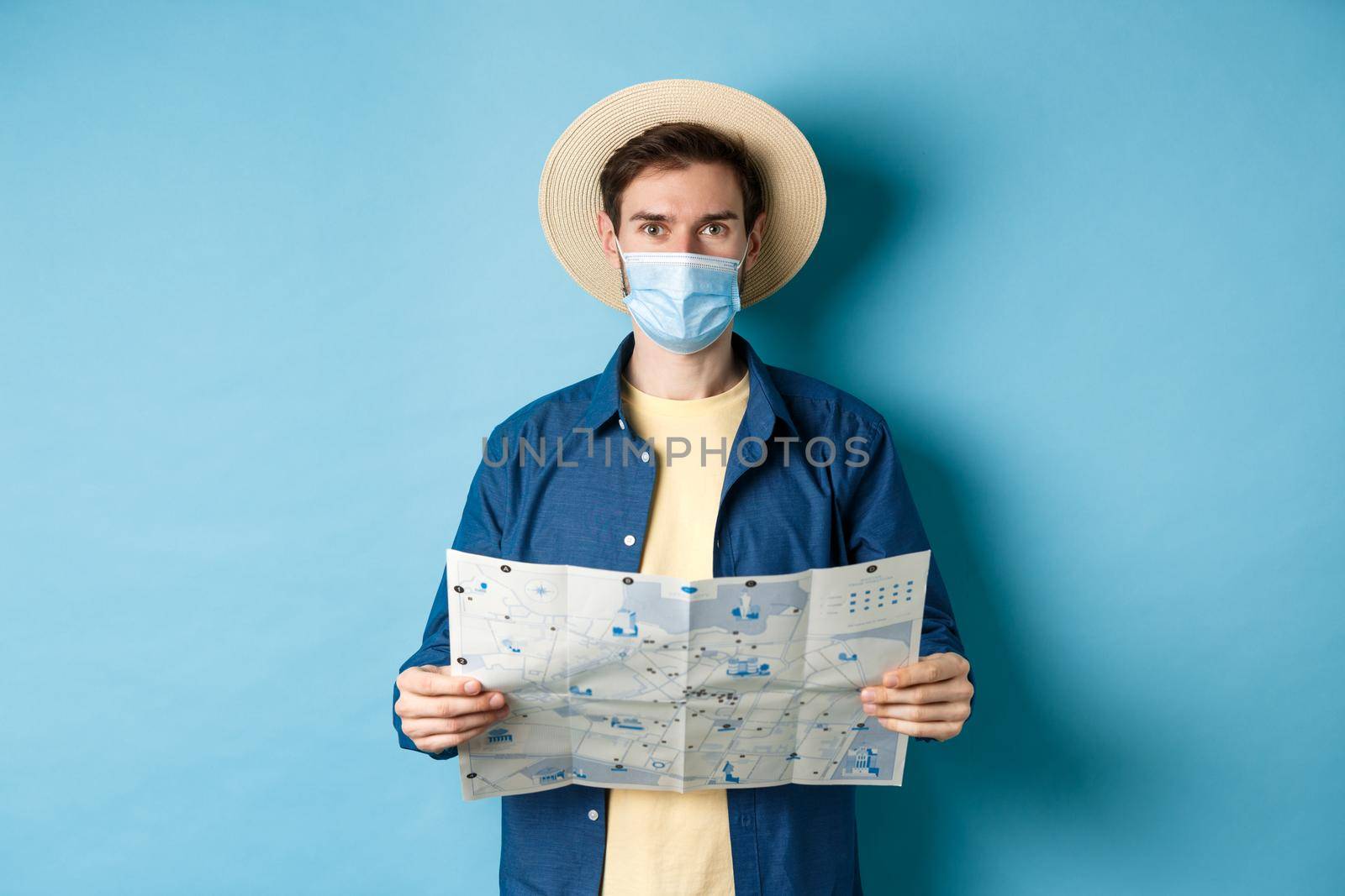 Covid-19, pandemic and travel concept. Happy tourist on summer vacation wearing medical mask and straw hat, holding road map, standing on blue background.