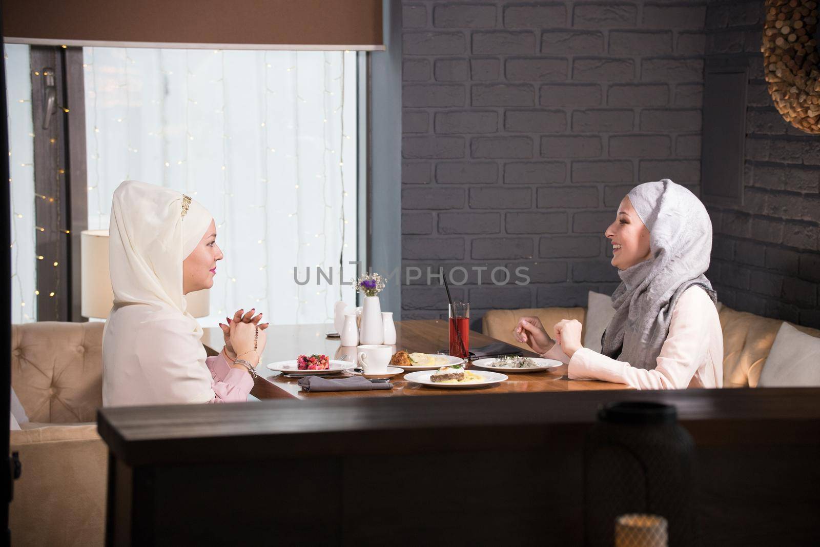 Two young Muslim women chatting at a table in a cafe, telephoto shot