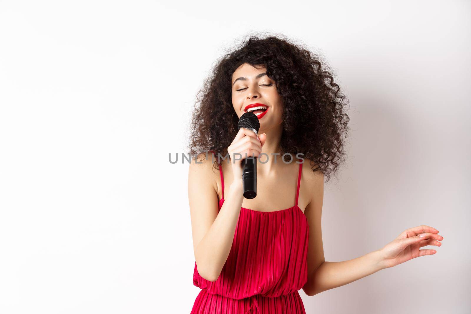 Happy elegant woman in red dress performing with microphone, singing karaoke and smiling, standing over white background.