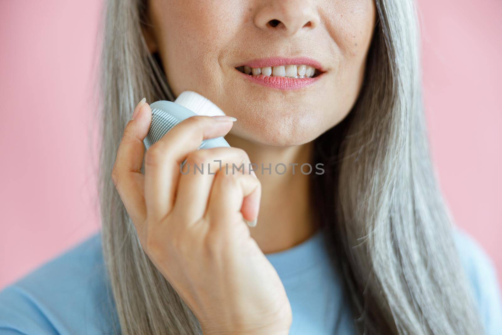 Happy middle aged grey haired woman cleans face with sonic brush standing on pink background in studio closeup. Mature beauty lifestyle