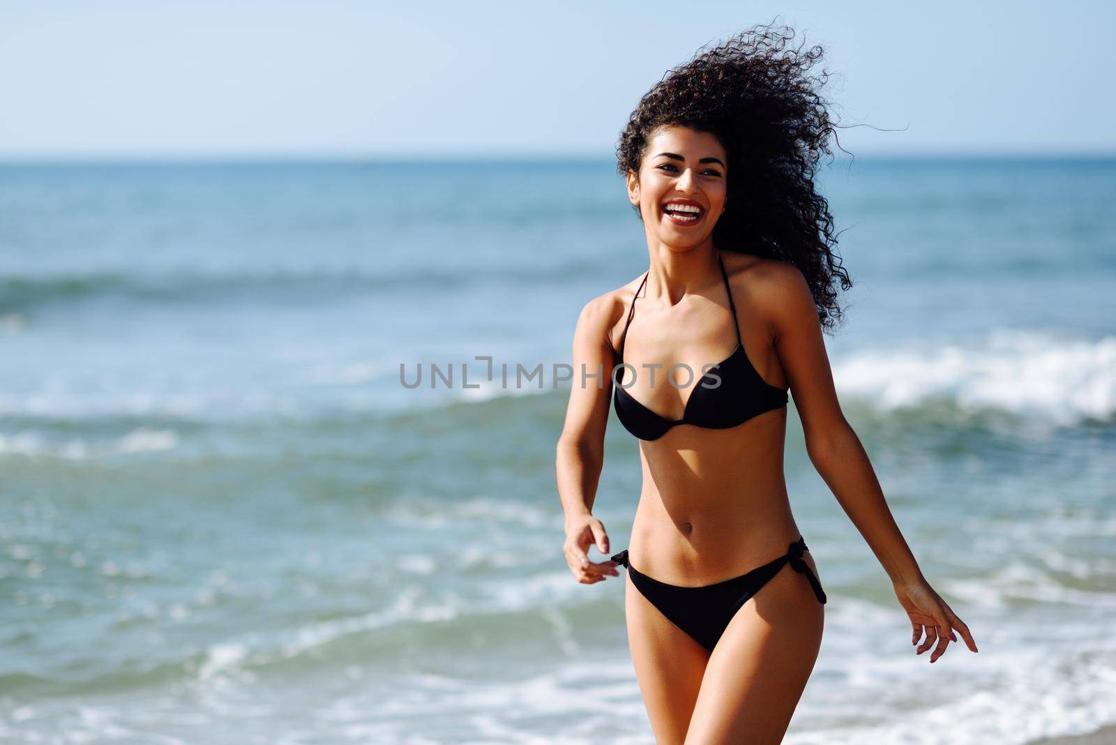 Young arabic woman with beautiful body in swimwear smiling in a tropical beach. Brunette female with curly long hairstyle wearing black bikini.