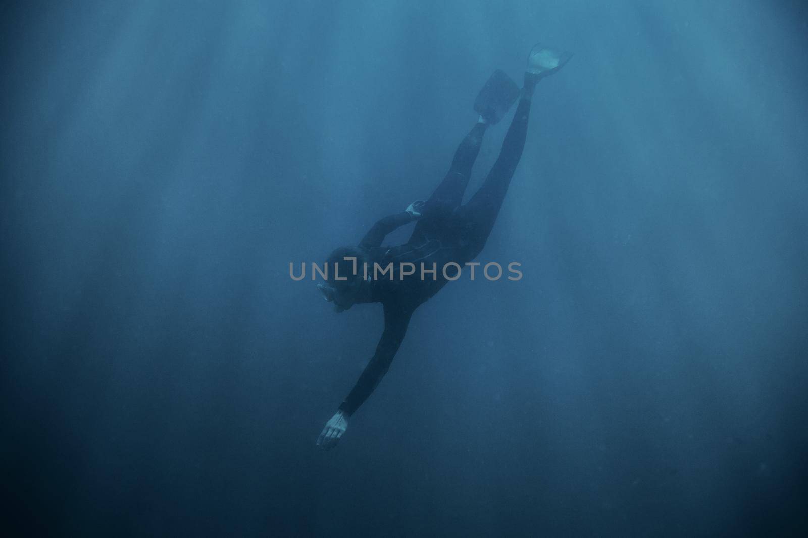 Sporty young man freediver wearing in wetsuit and flippers diving underwater in blue deep sea.