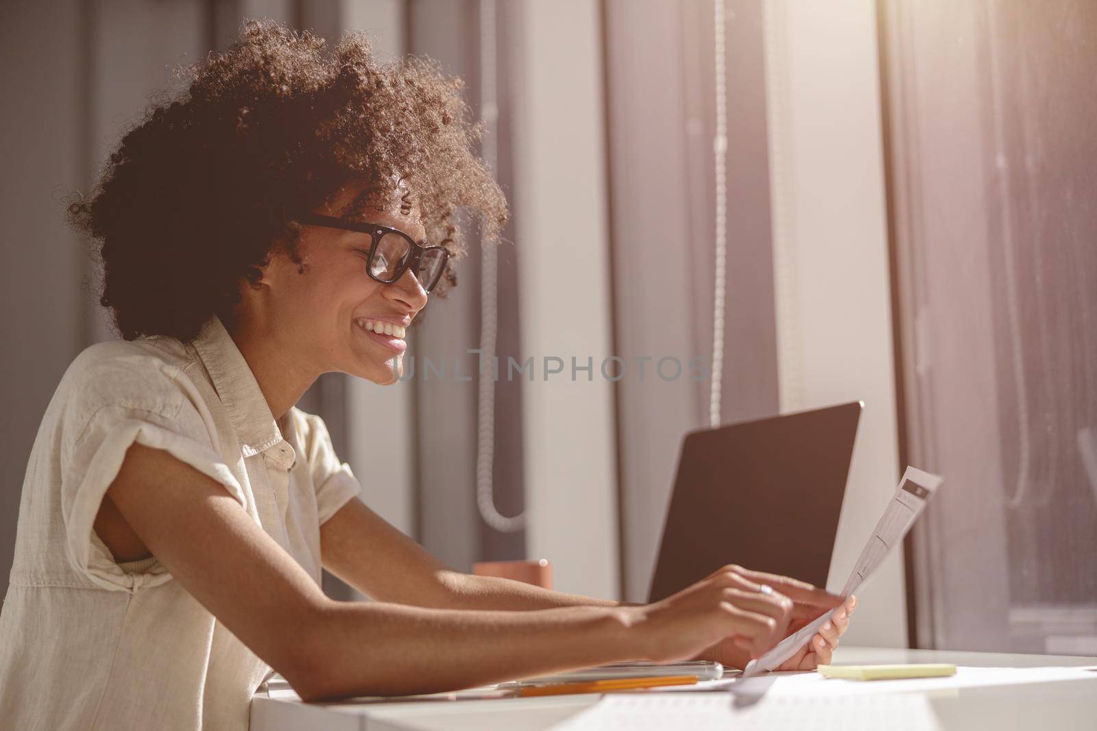 Happy pretty female worker working with laptop while holding document and sitting near the window