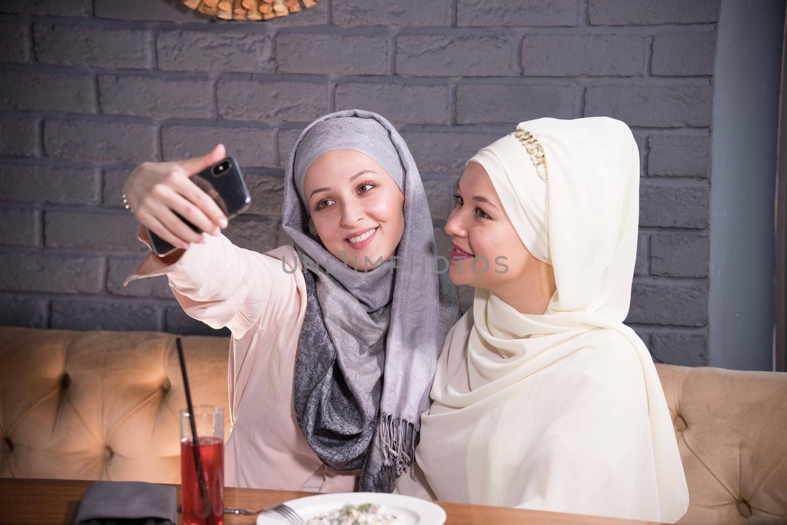 Two young Muslim women taking selfie in a cafe, telephoto shot