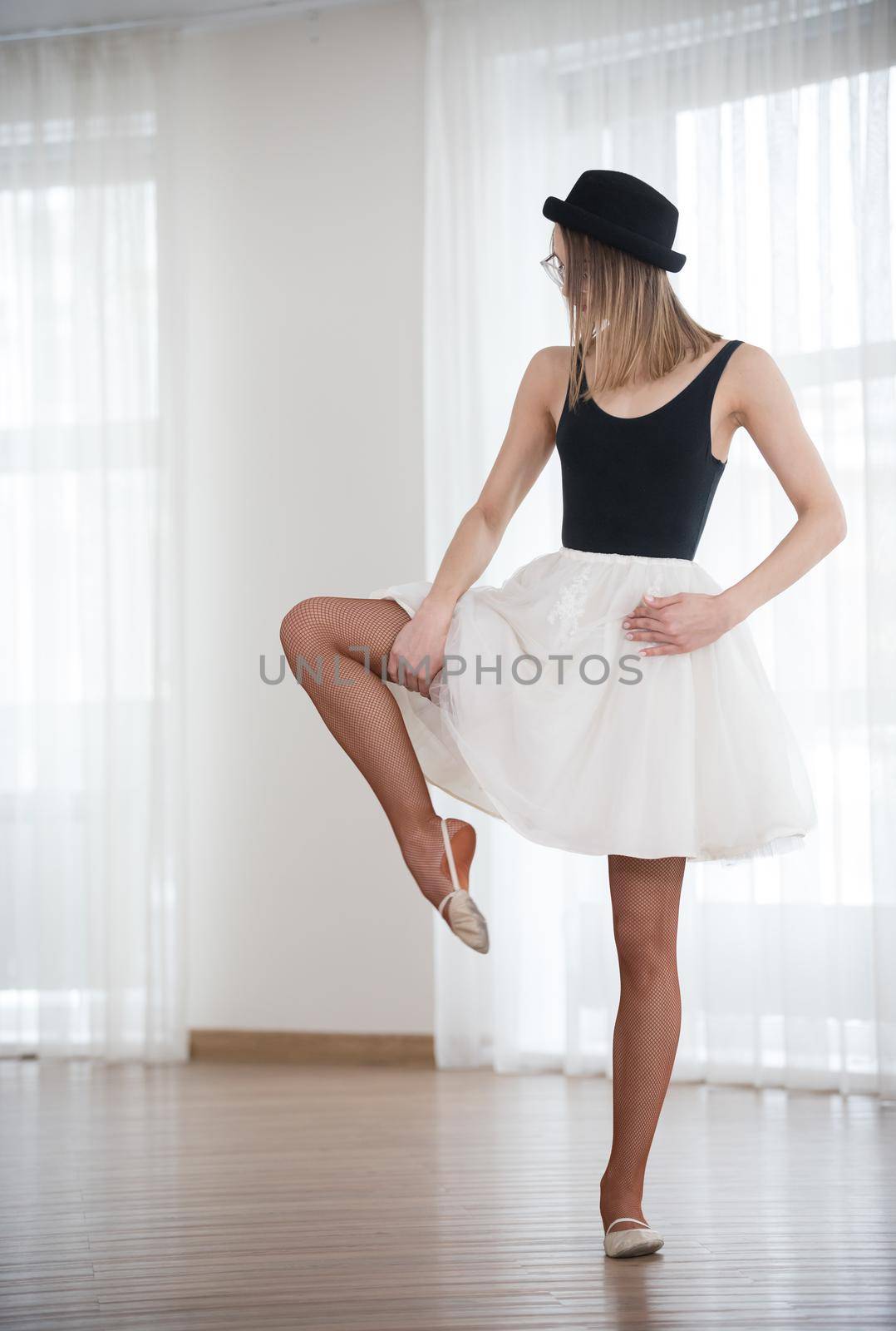 Ballet girl in a hat bends a leg to the side, in the studio, telephoto shot