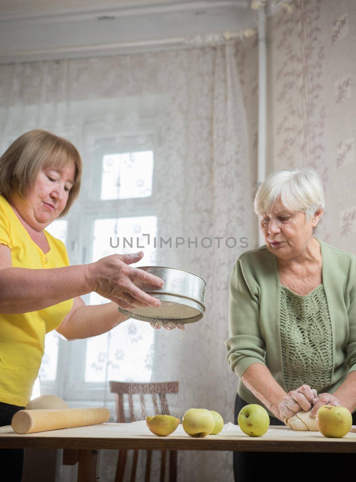 Two elderly woman making apple little pies at home. Sifting flour on the table. Mid shot