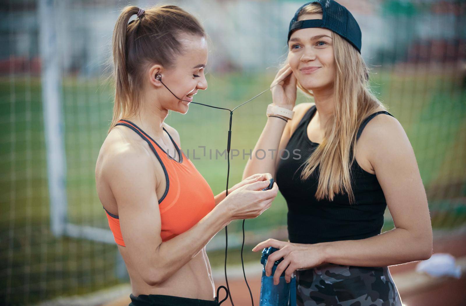 Two young athletic woman resting up after training. Listen music in headphones. Mid shot