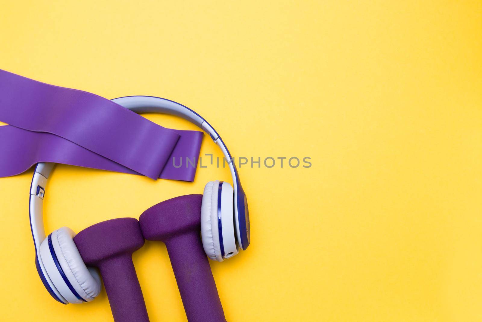 Sports equipment,gray and blue headphones and purple dumbbells on a yellow background, purple rubber expanders, copy space, top view, sport and music concept