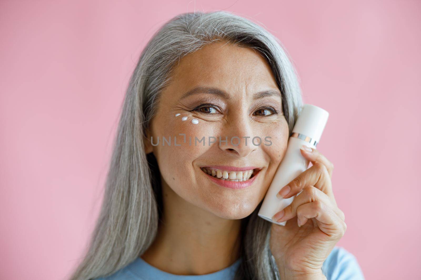 Smiling middle aged Asian woman with cream drops under eye holds bottle near face on pink background in studio. Mature beauty lifestyle