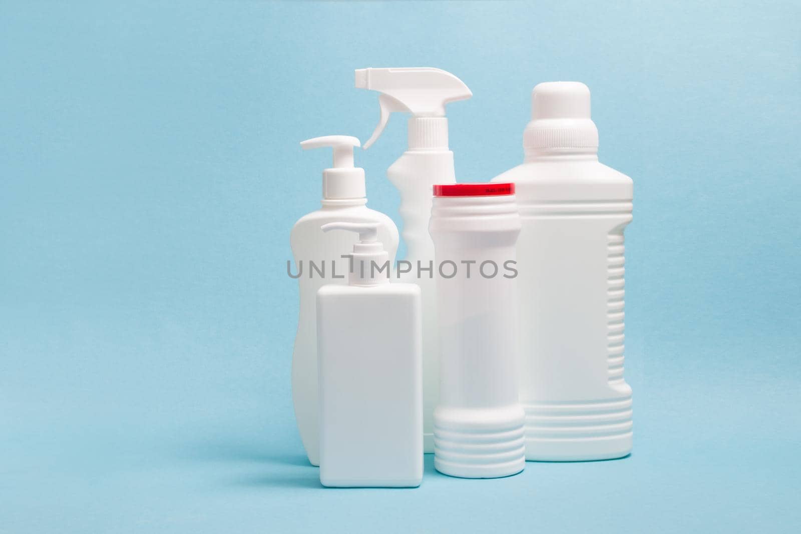 several white plastic bottles without labels with cleaning products on a blue background, washing gel, two bottles with dispensers, a spray bottle and a bottle with cleaning powder
