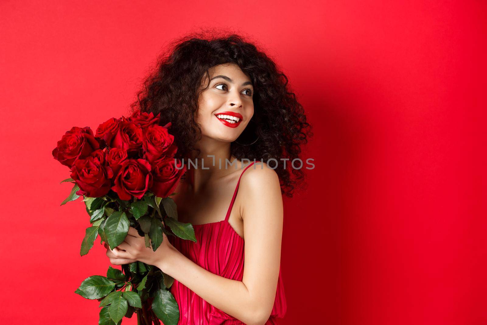Tender young woman in elegant red dress, holding romantic bouquet of red roses and looking right, smiling dreamy, thinking about lover on Valentines day, studio background.