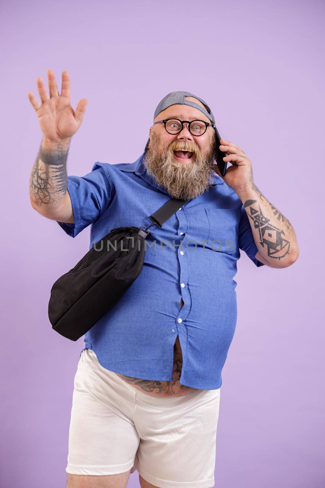 Jouful plump man talks by phone and waves hand standing on purple background by Yaroslav_astakhov