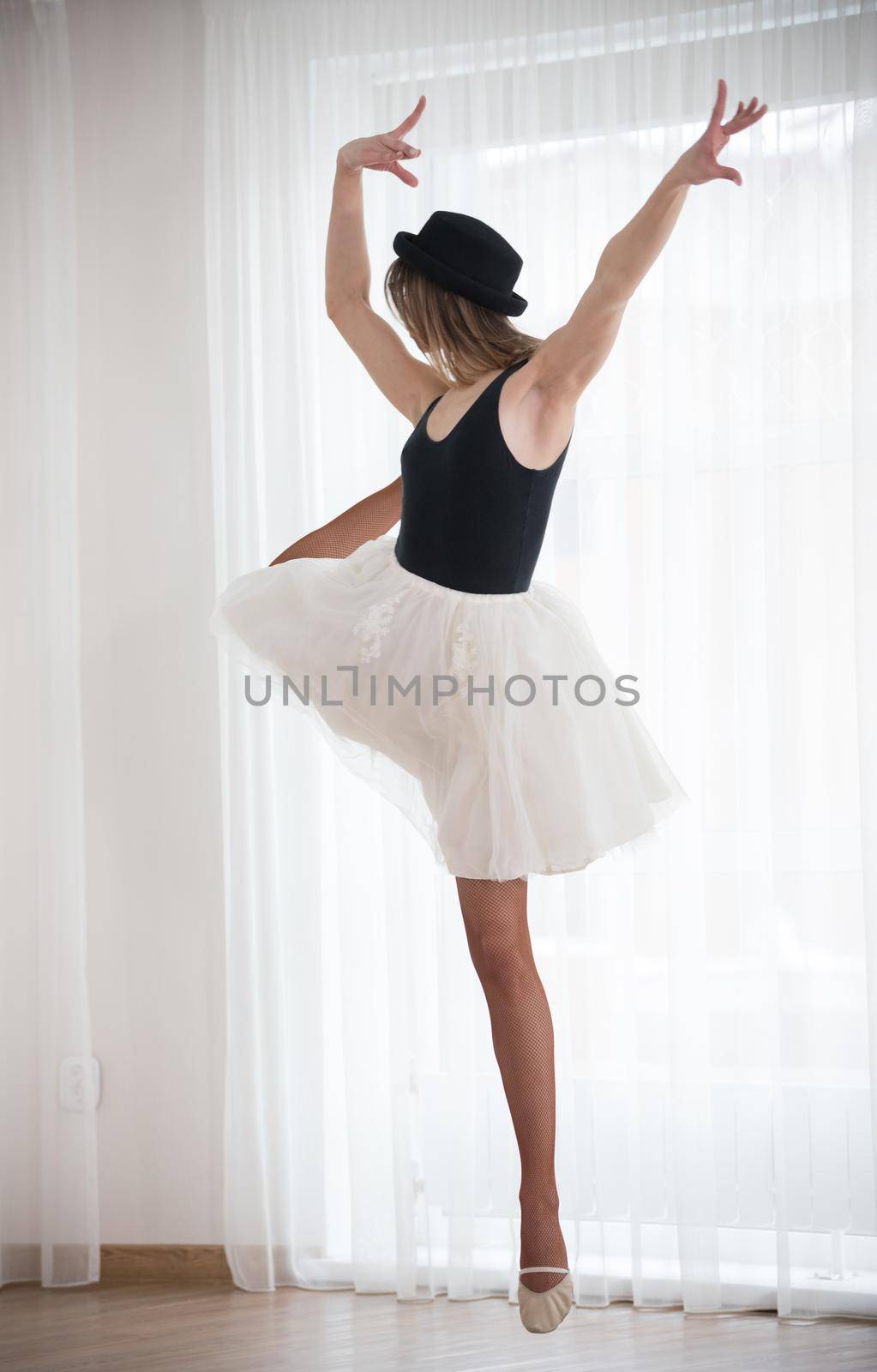Ballerina in hat stands near the window, raises her leg, in a bright studio, telephoto shot