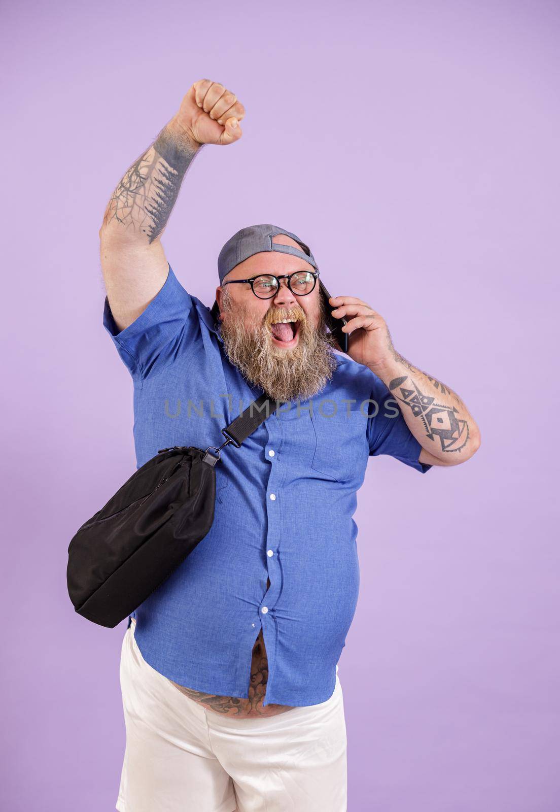 Excited middle aged obese man with crossbody bag talks on smartphone raising up fist on purple background in studio