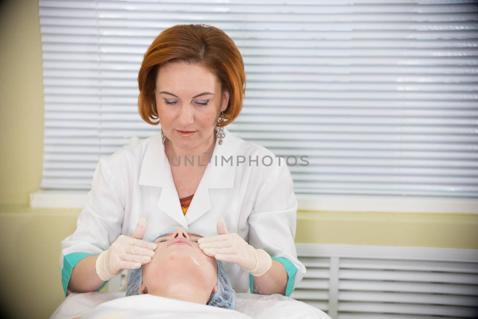 Doctor with red hair doing a facial massage to a young woman and talking to her during the procedure, front view