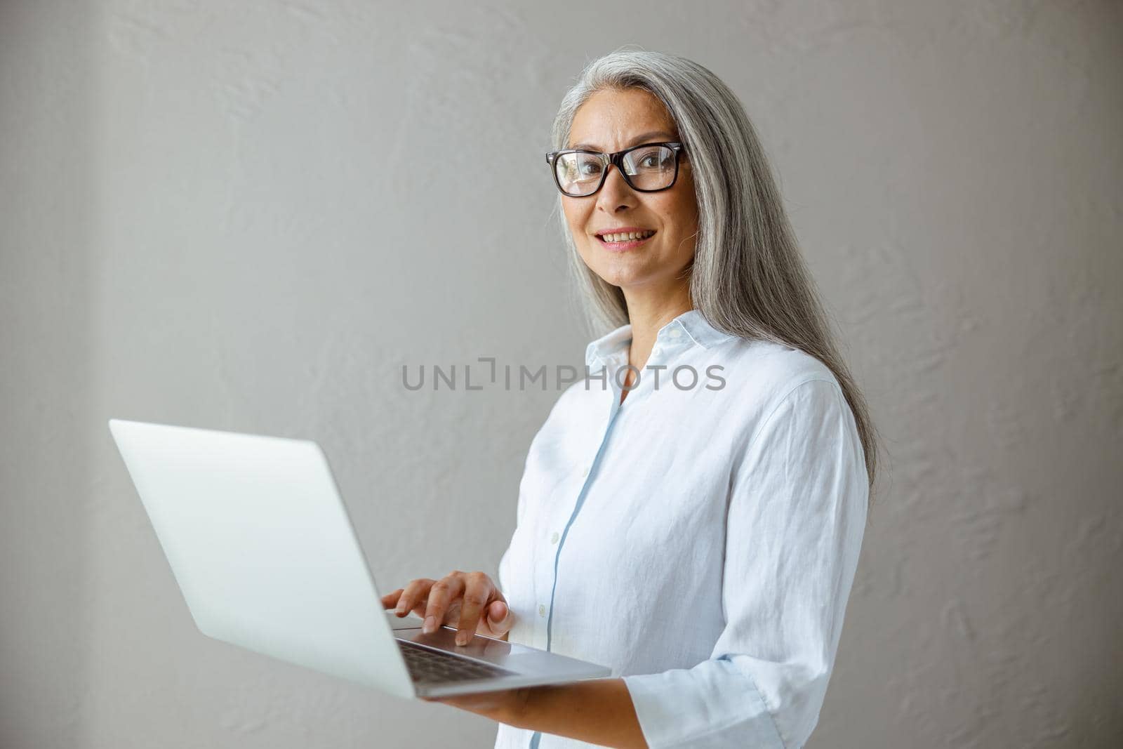 Positive hoary haired middle aged Asian woman in white blouse touches pad of laptop standing near grey stone wall in studio
