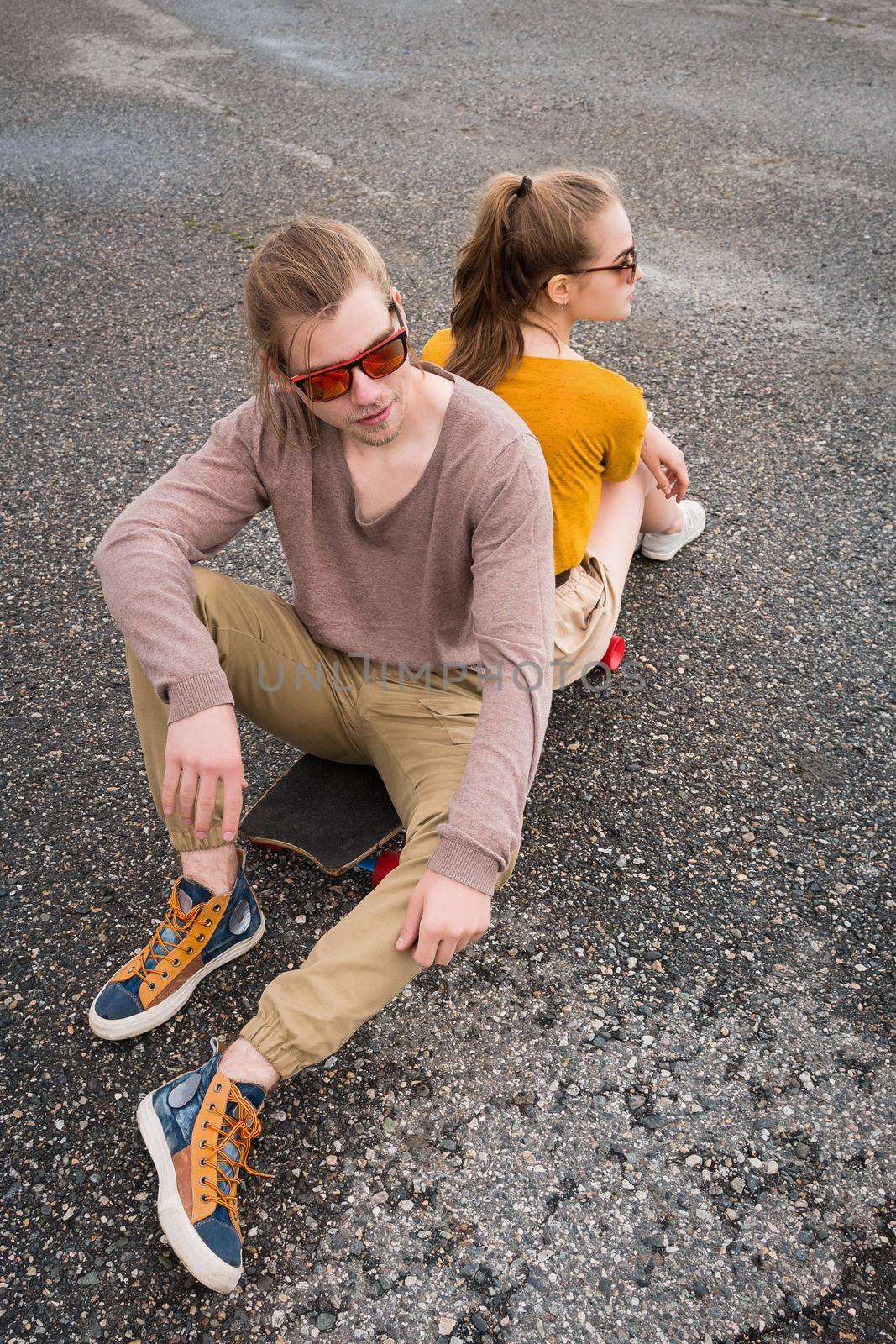 An attractive young couple in sunglasses sit back to back on their longboard in a suburban parking. The concept of a young family generation millennials style and leisure.