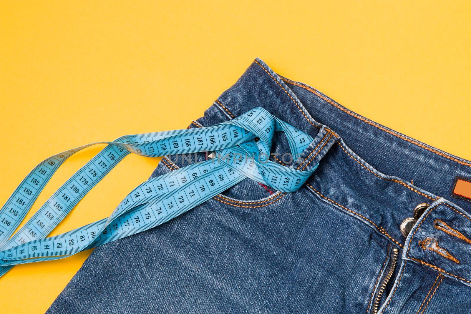 blue measuring tape is threaded into jeans instead of a belt, blue jeans on a yellow background, copy space, weight loss and weight control concept, measuring body volume