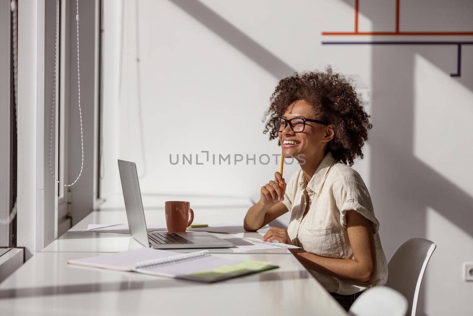 Happy pretty multiethnic lady in glasses sitting at desk while working with laptop and documents