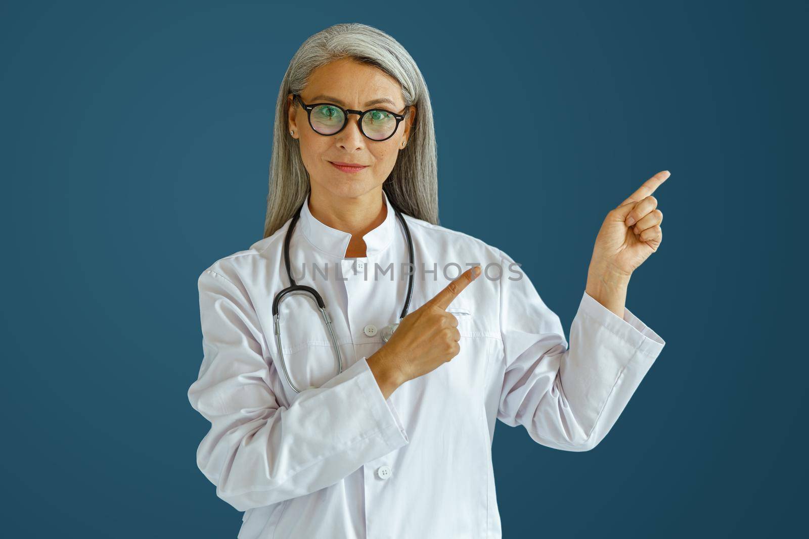 Smiling middle aged woman doctor in white uniform with glasses points aside posing on blue background in studio. Professional medical staff