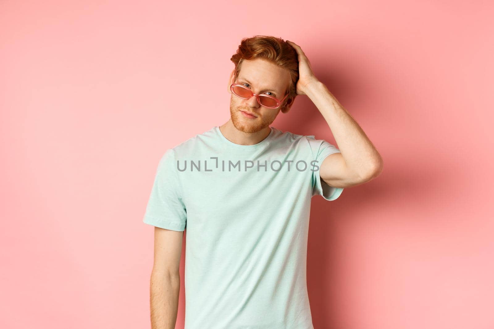 Handsome young redhead man in sunglasses, brushing hair with hand and looking smug and confident at camera, standing over pink background.