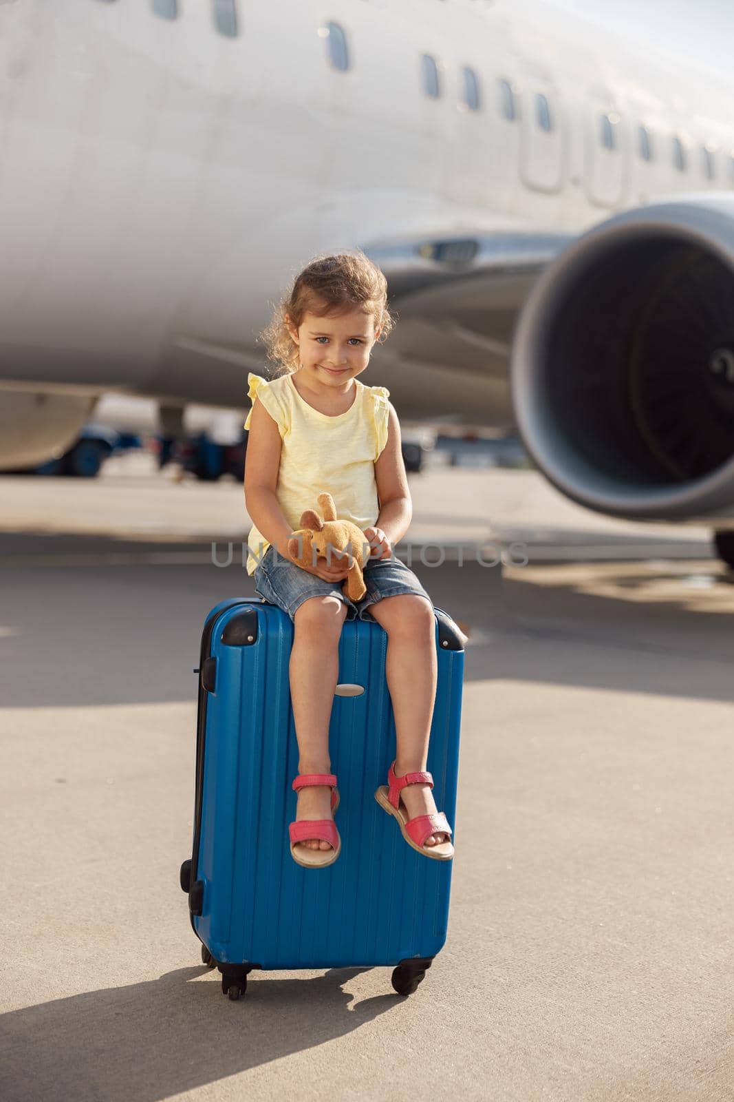 Full length shot of cute little girl holding her toy, looking at camera and sitting on suitcase in front of big airplane by Yaroslav_astakhov