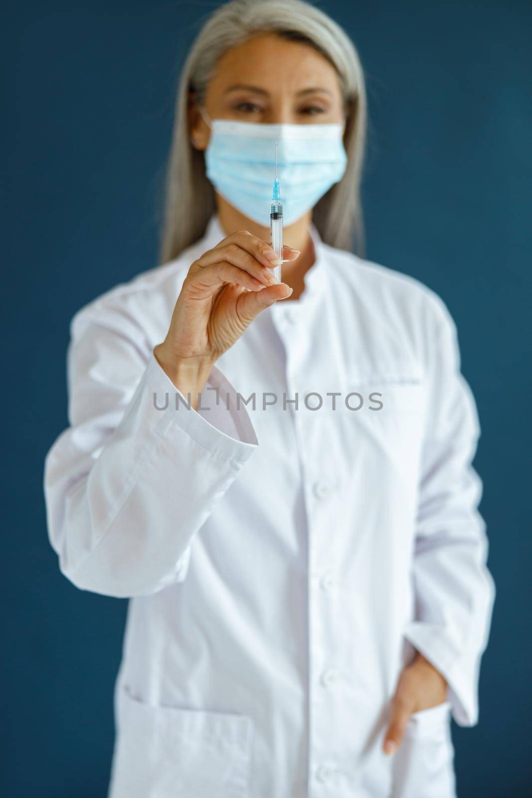 Middle aged Asian lady doctor in white robe with medical mask stands on blue background in studio, focus on hand with syringe. Vaccination day