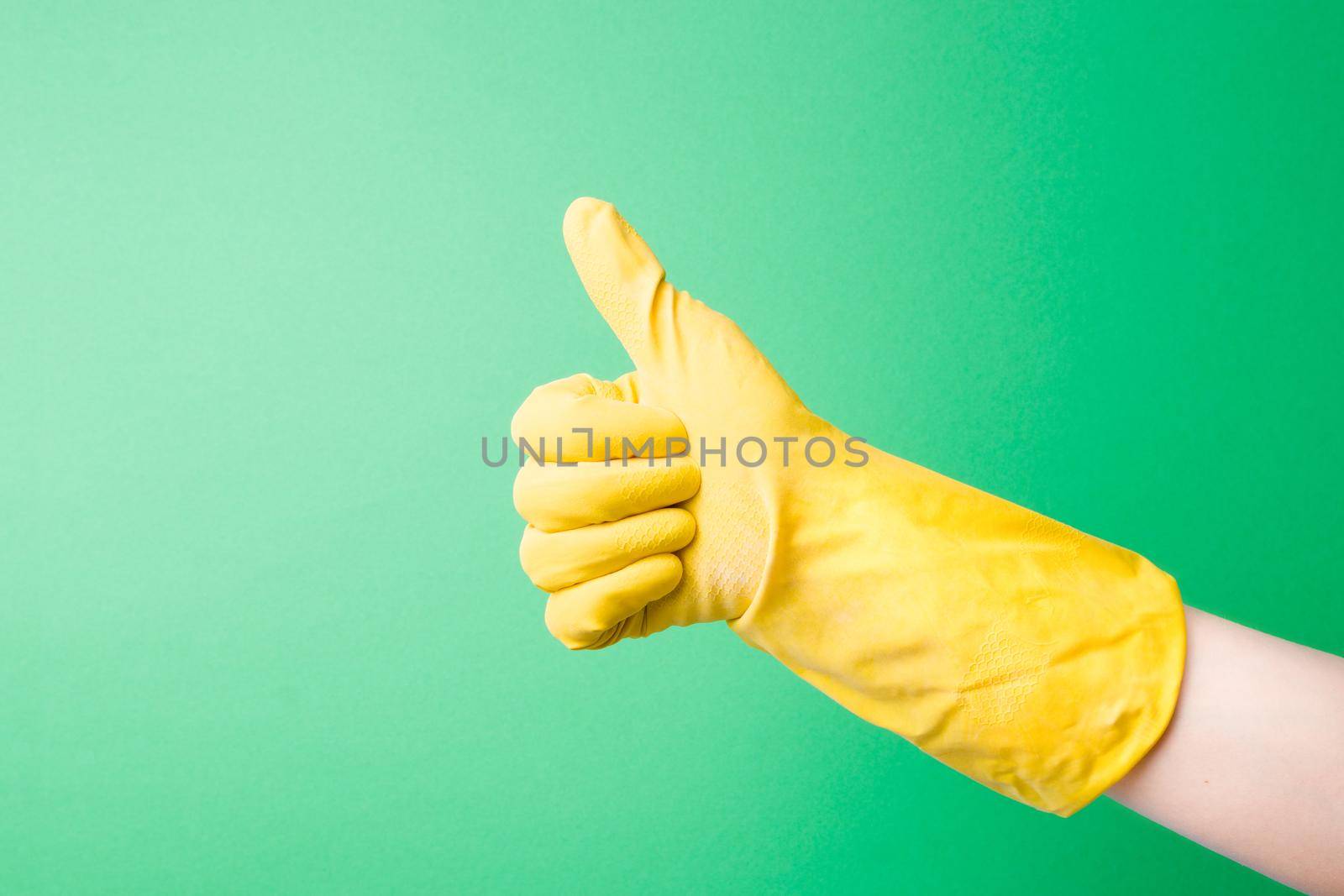 female hand in yellow rubber glove shows thumbs up gesture, seal of approval, cleaning or construction work is completed successfully, green background copy space, cleaning company and spring cleaning concept