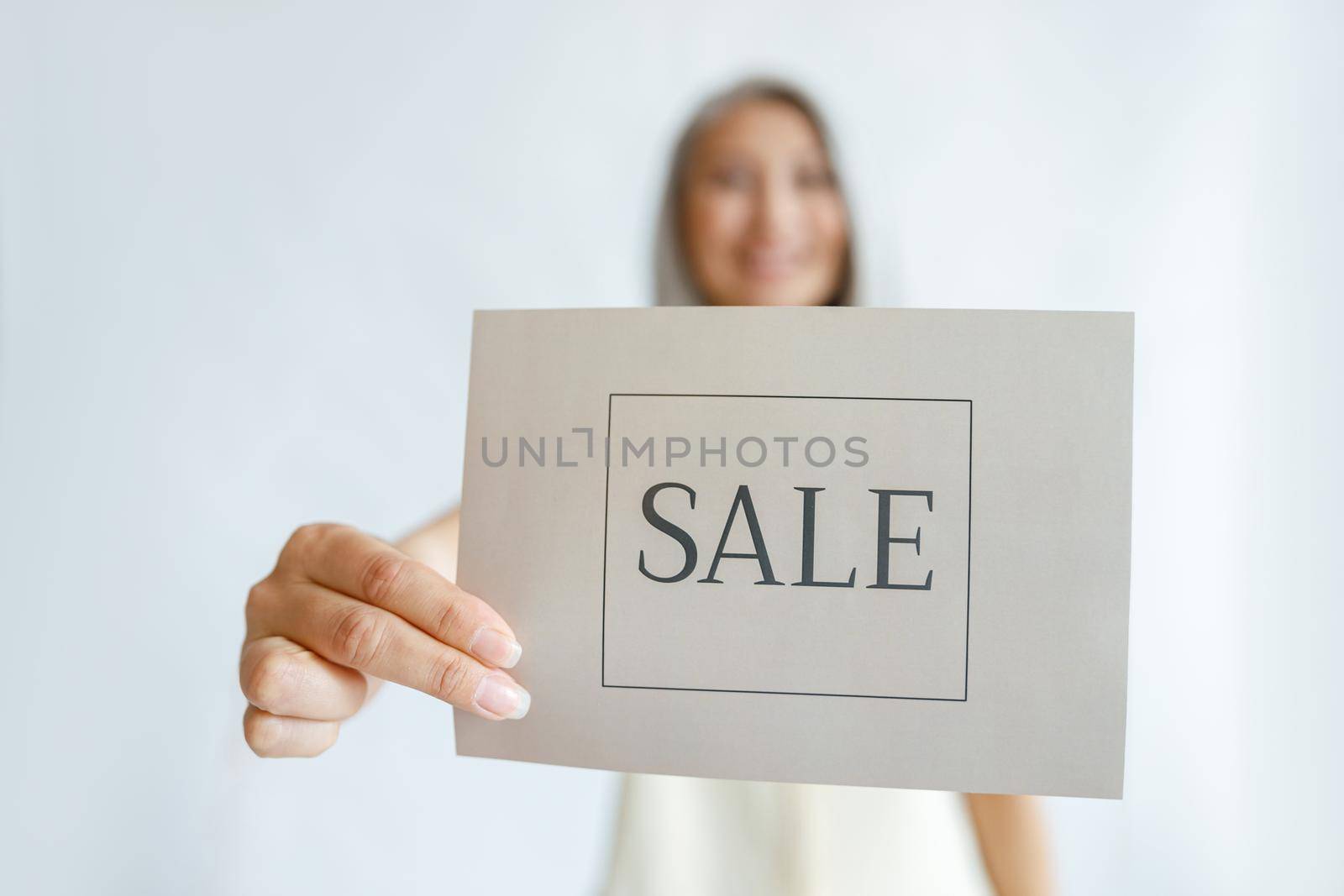 Positive mature woman stands on light background in studio, focus on hand holding card with word Sale closeup